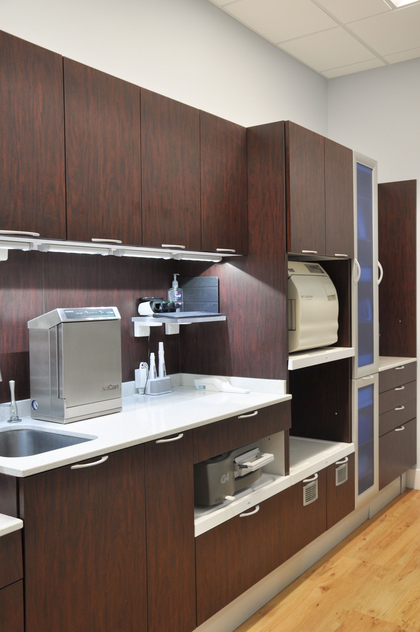 Dark wood cabinetry in a dental office. Stainless steel sterilizers and sink against a white countertop.