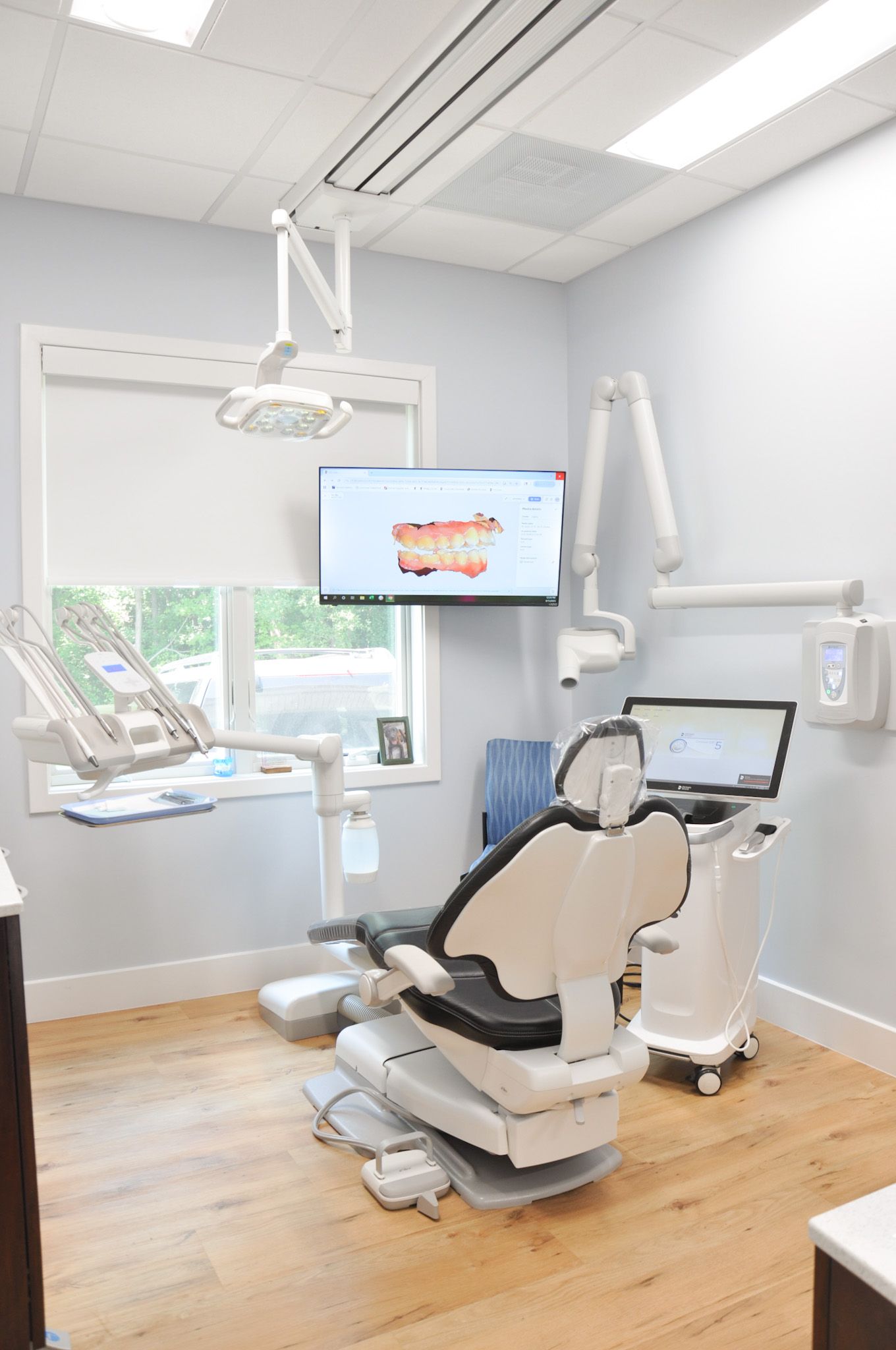 Dental office with chair, monitor, and tools, lit by overhead lighting, wood floors, and a window.