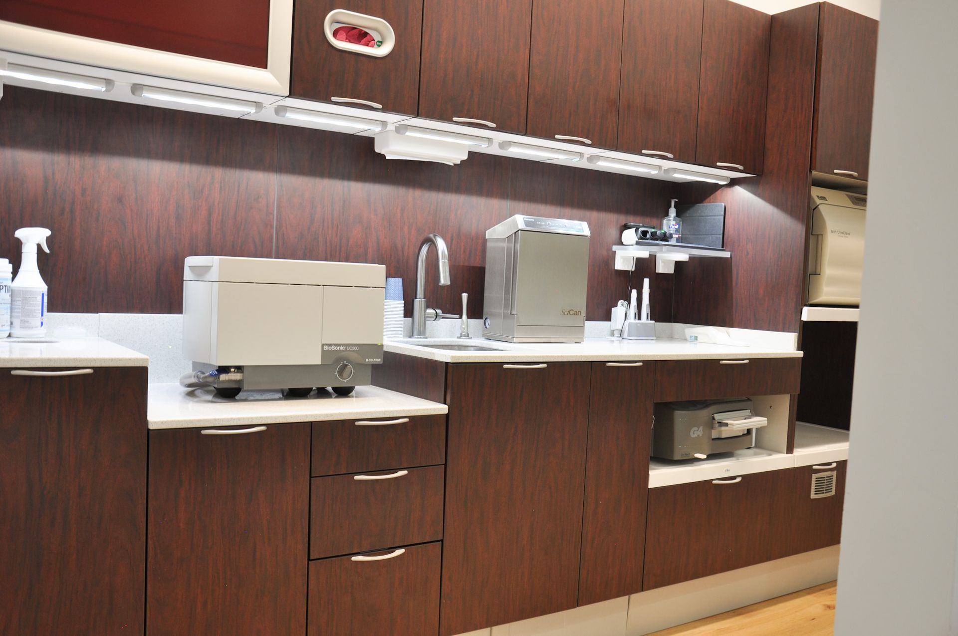 Dental office cabinetry with equipment: a sink, sterilizer, and storage against dark wood-grain cabinets.