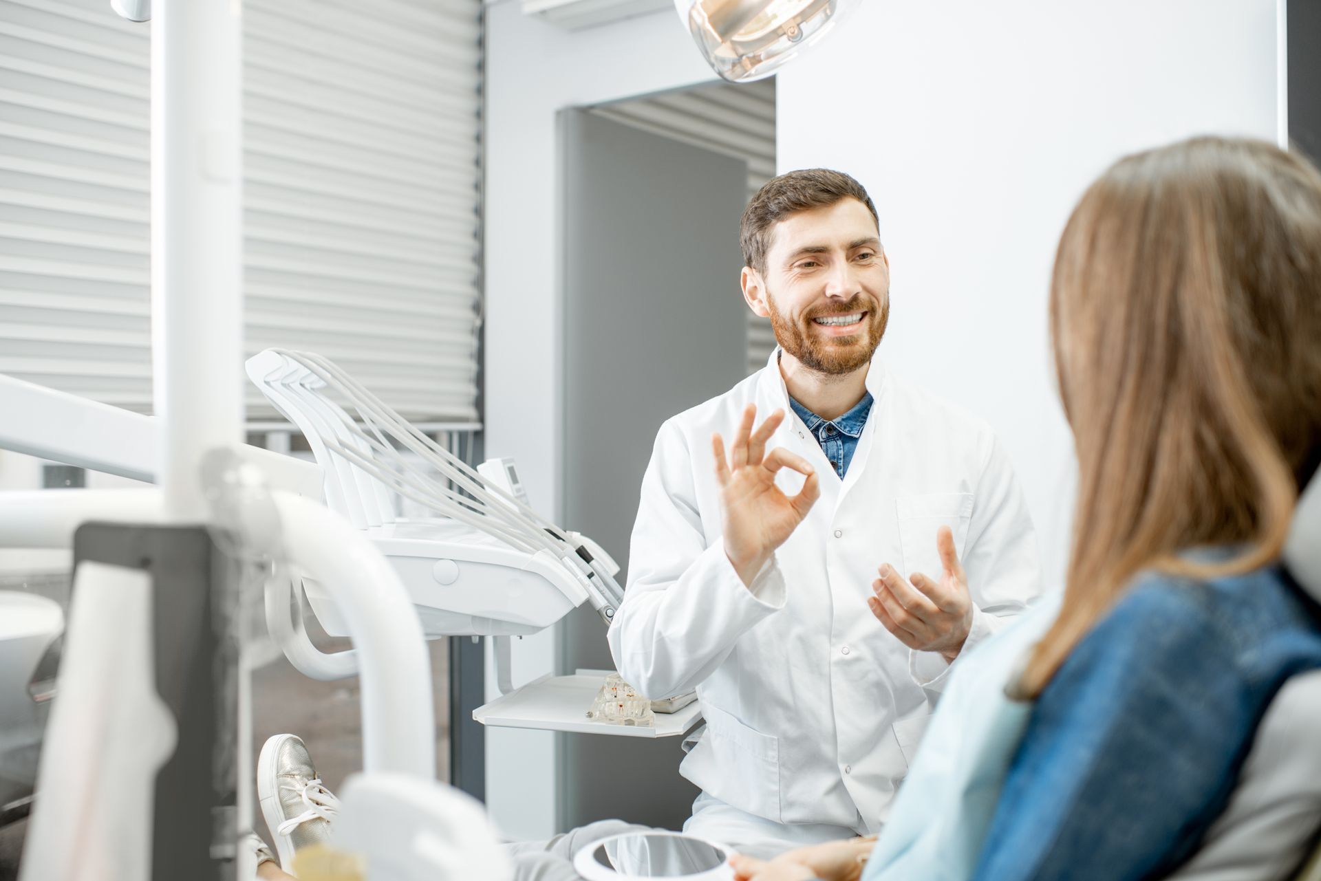 Dentist in white coat explains procedure to patient in dental chair, smiling, gesturing