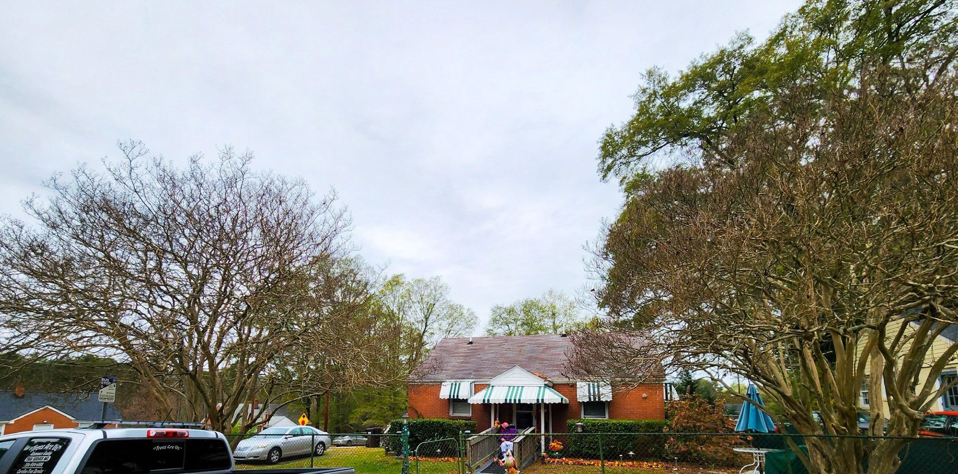A red-brick house with a peaked roof and an awning, flanked by large trees under a cloudy sky.