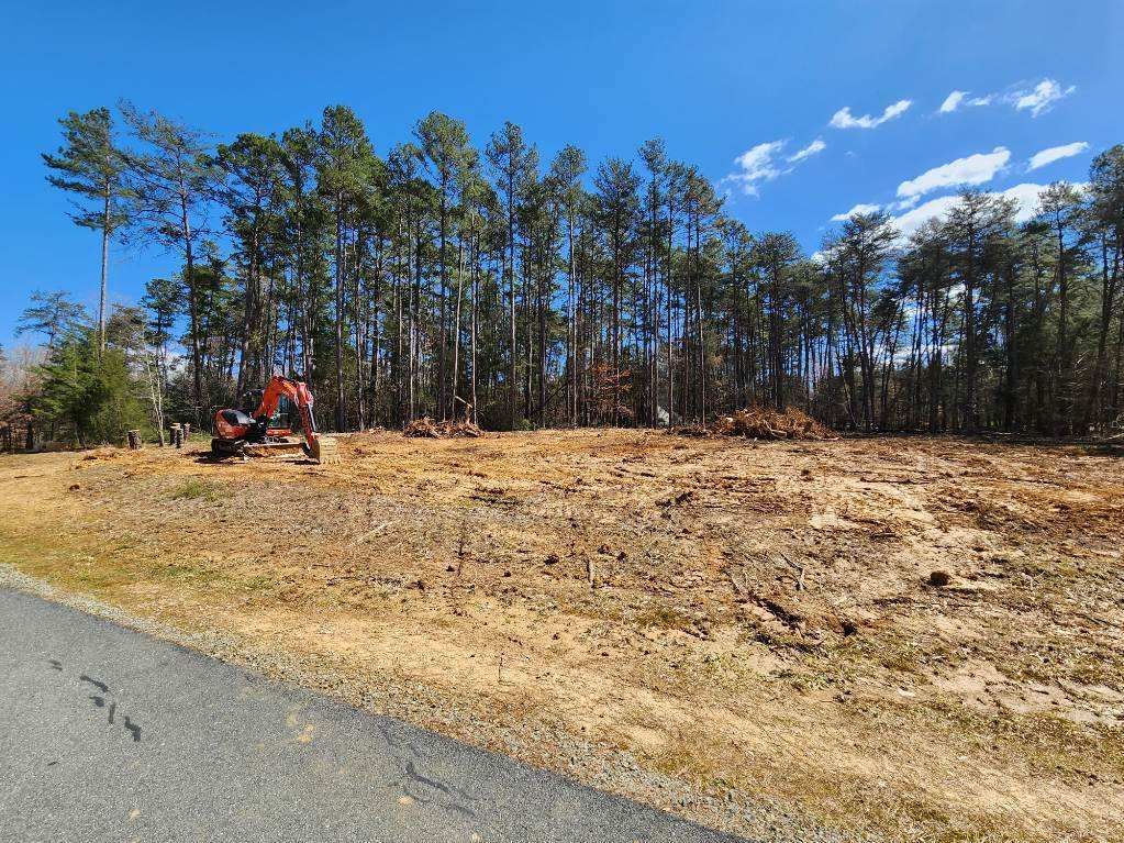 An orange excavator works on a cleared field of dry, brown grass in front of a dense line of pine trees under a blue sky.