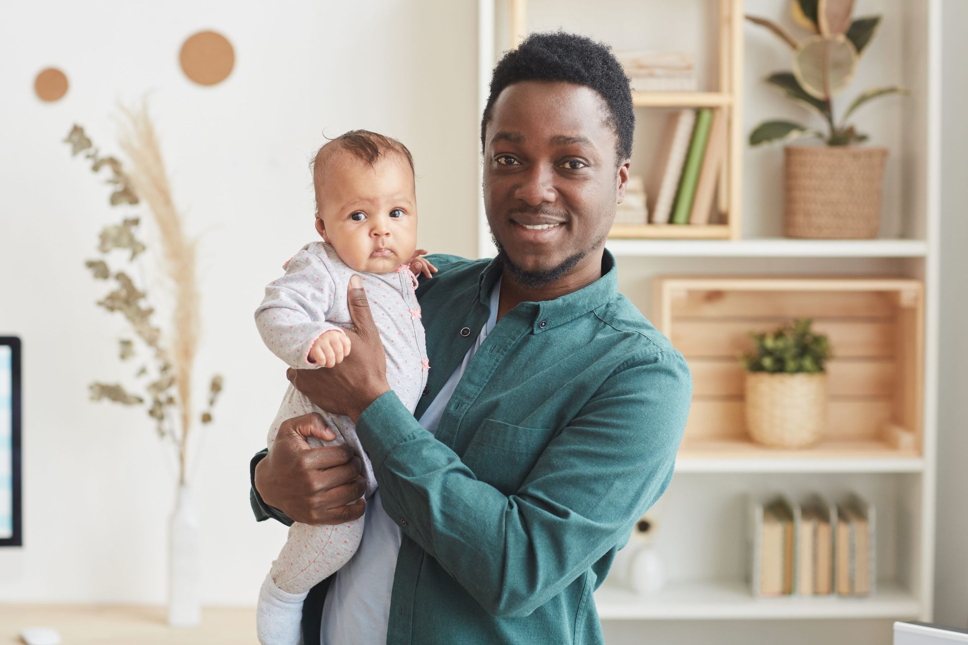 Person holding a baby in a bright living room, both facing the camera and smiling.