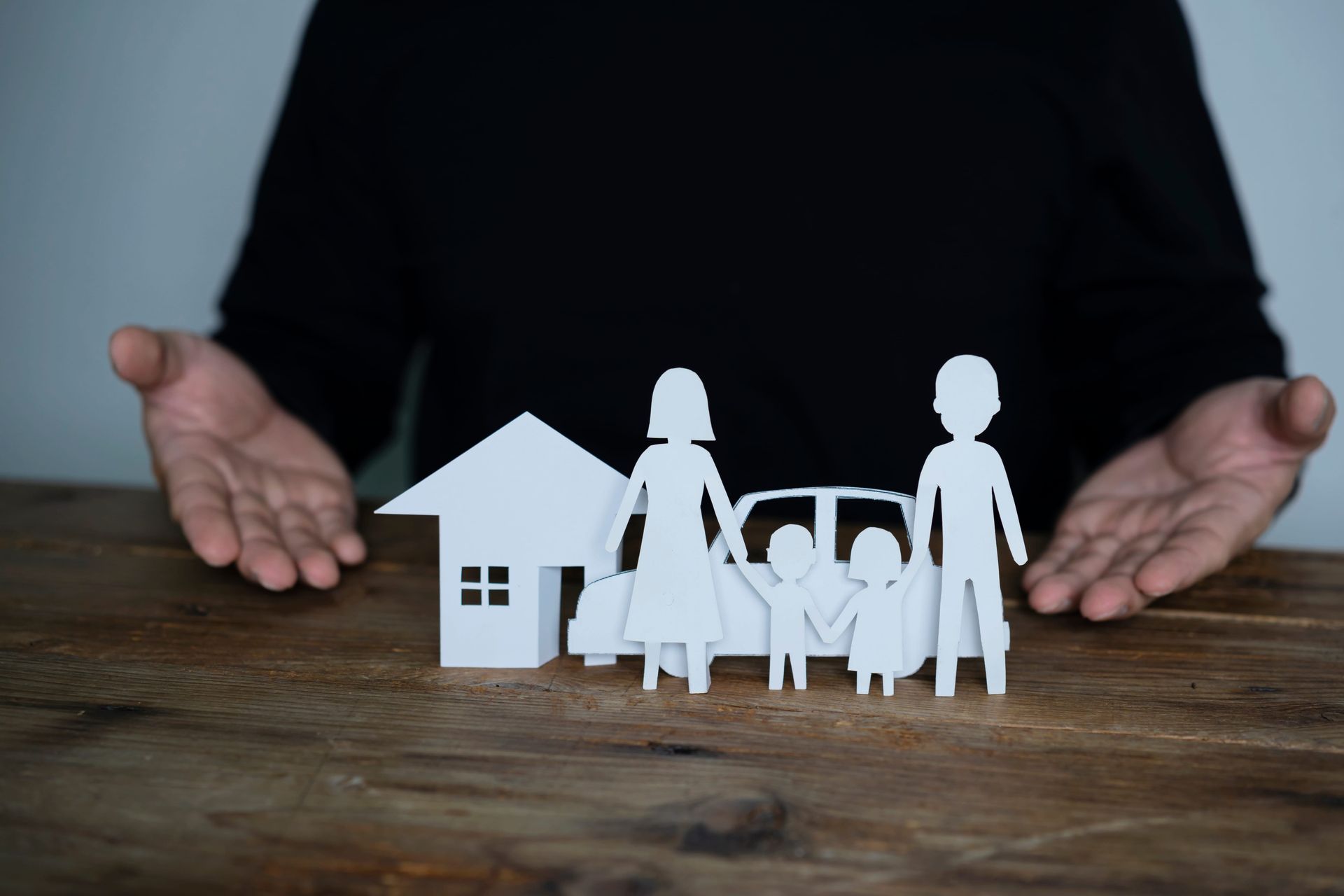Paper family figures in front of a house on a wooden table, with hands framing them in the background