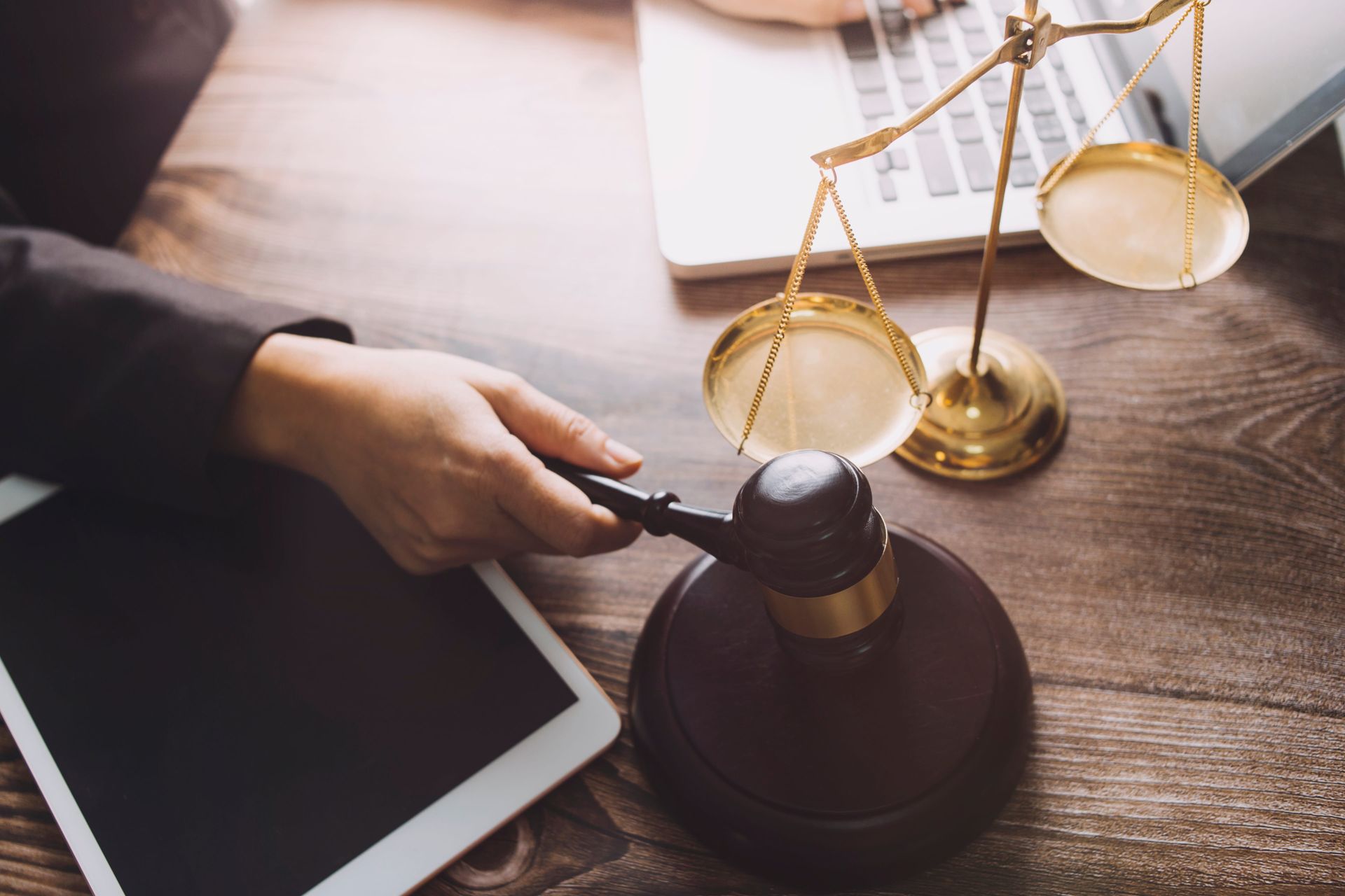 Lawyer’s hand holding a gavel beside scales of justice and a tablet on a desk