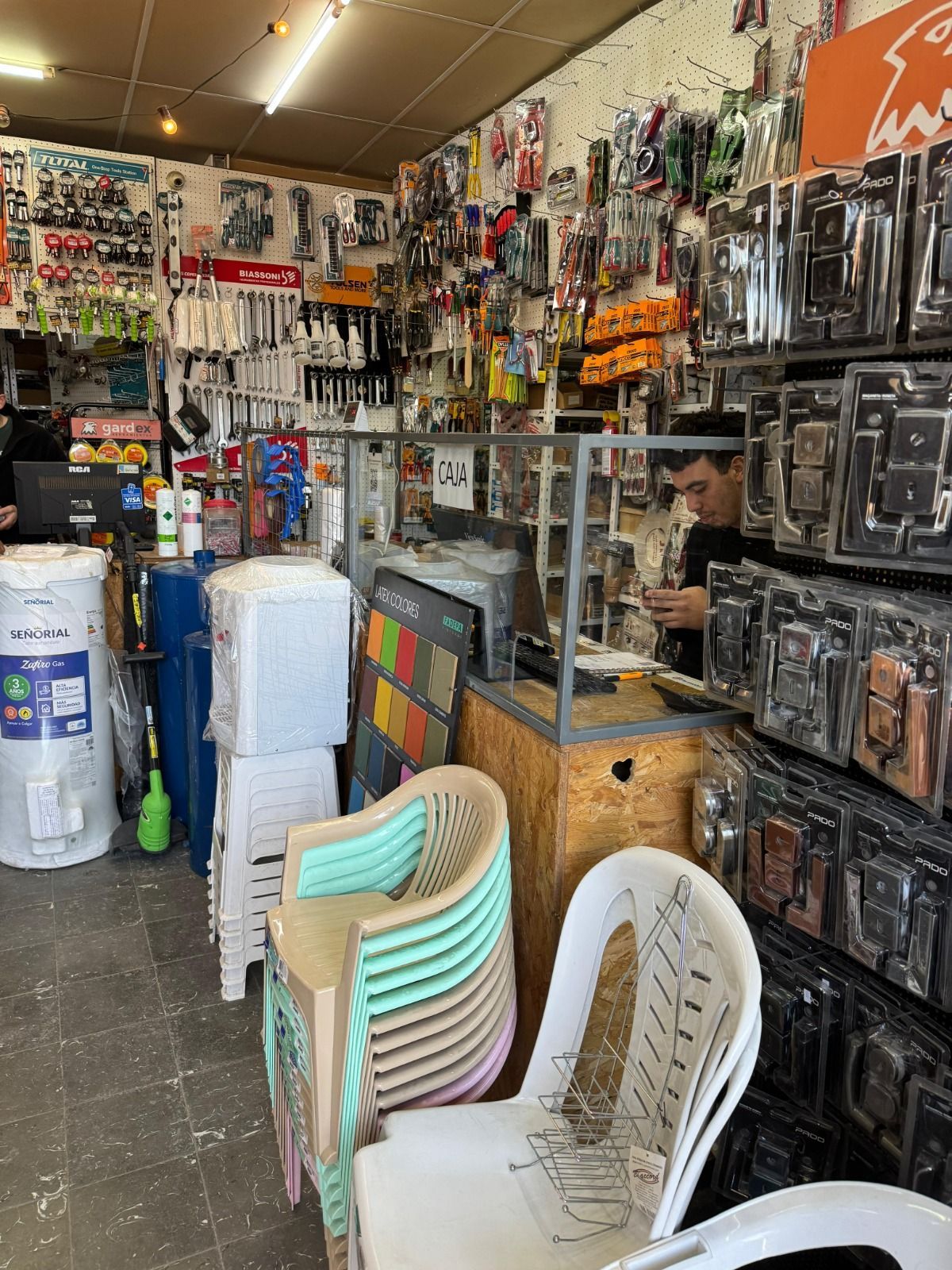 El interior de una ferretería desordenada. Una persona trabaja tras un mostrador; la mercancía se alinea en las paredes. Hay pilas de sillas en primer plano.