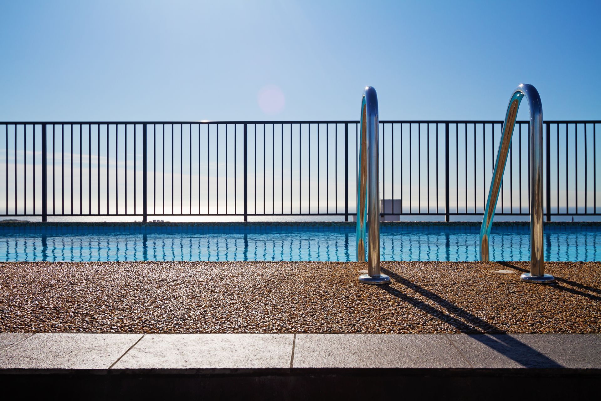 A swimming pool edge with a ladder and a fence and a blue sky in the background A swimming pool edge with a ladder and a fence and a blue sky in the background