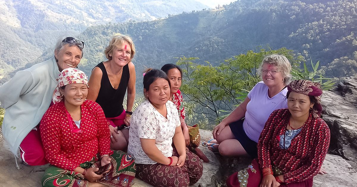 Four local women and three tourist women sit outdoors on a hillside, enjoying the view while on the Millennium Trek in Nepal.