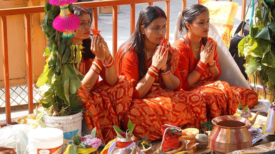 Three women in red traditional attire participate in a Nepali ceremony, hands in prayer, surrounded by offerings.