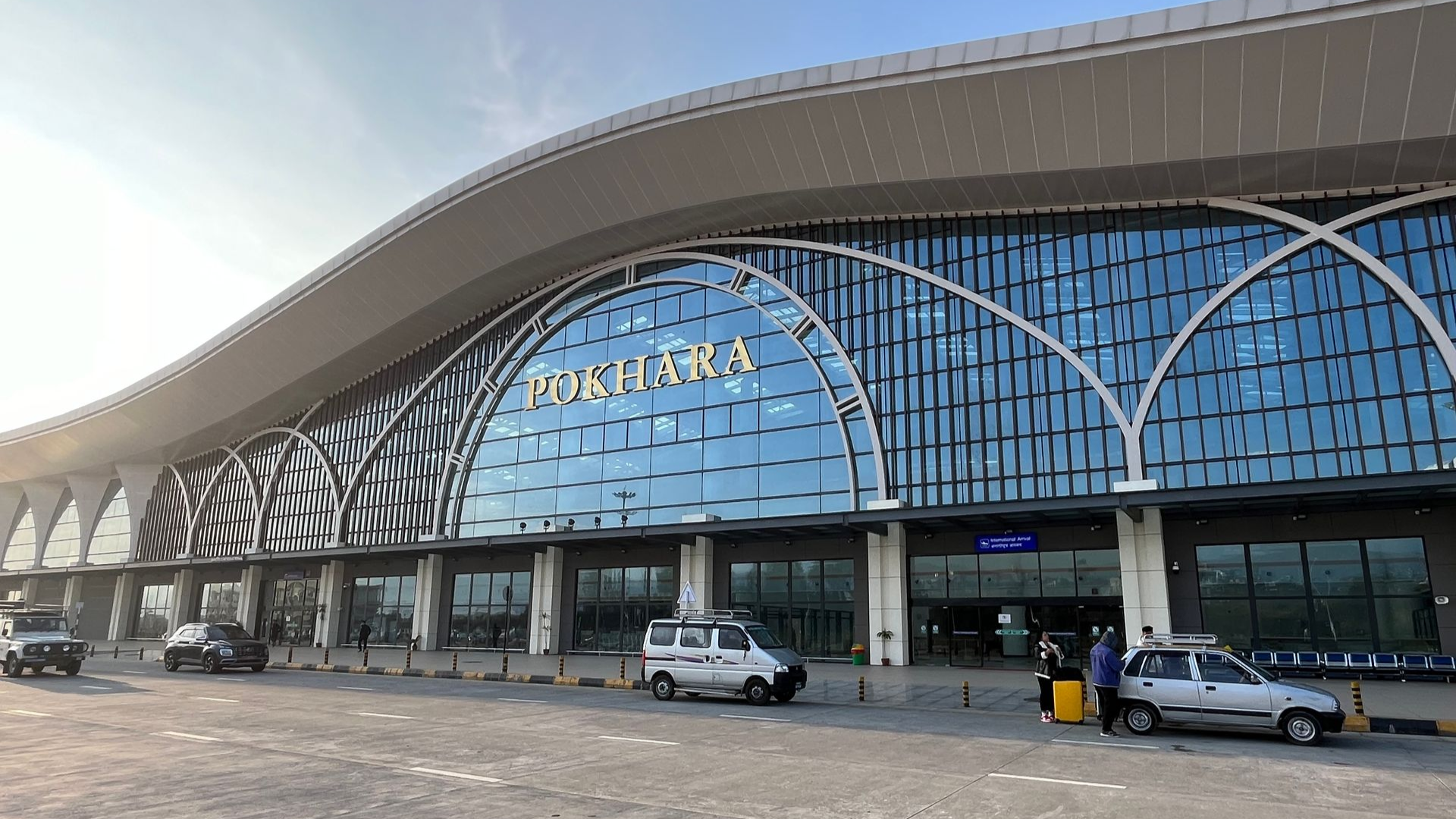 Pokhara International Airport's glass terminal with a wave-shaped roof, cars parked in front.