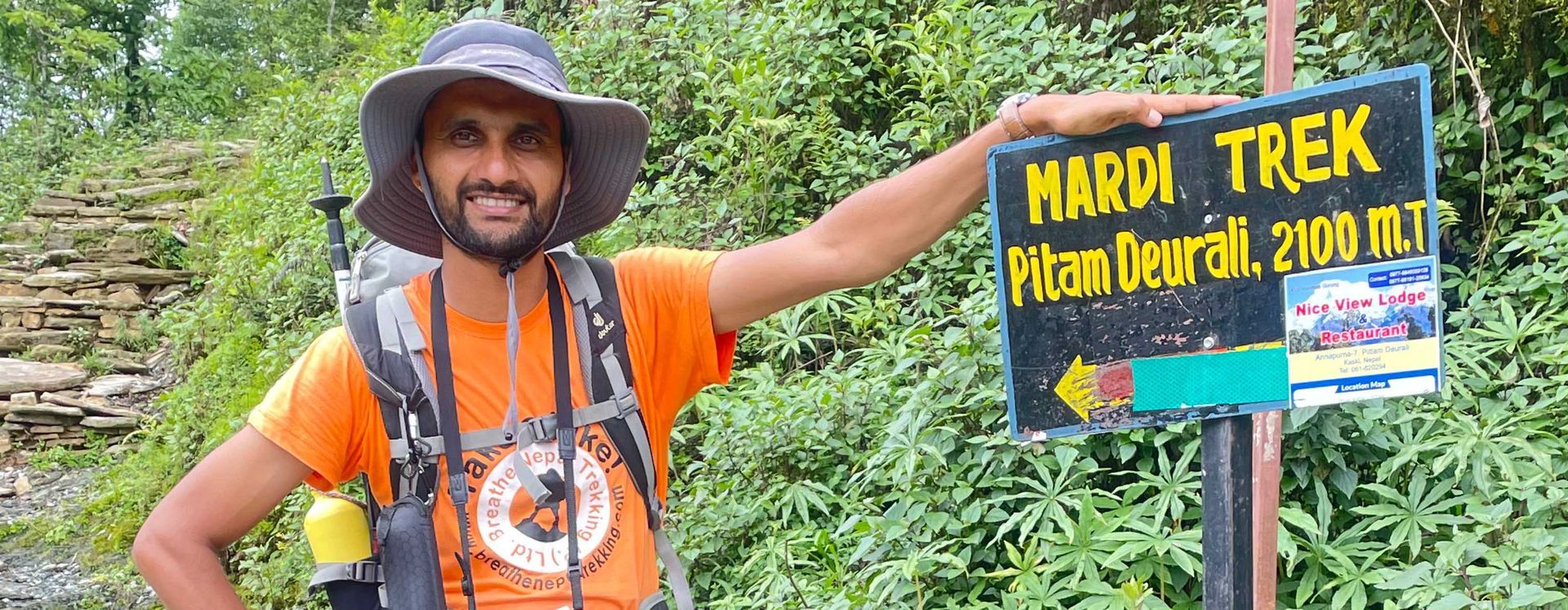 Trekking guide standing next to a Mardi Himal Trek sign, pointing towards the trailhead while surrounded by lush greenery.