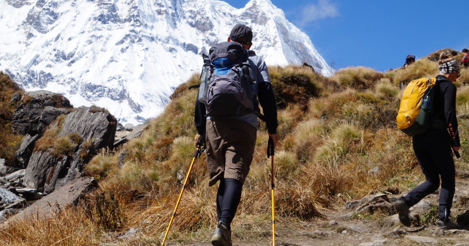 Trekkers hiking in the Annapurna region, Nepal, carrying backpacks and trekking poles with a snow-covered mountain ahead.
