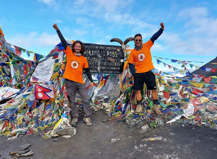 Two trekkers celebrate at Thorong La Pass with colorful prayer flags behind them, showcasing their achievement.