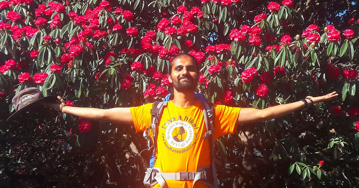 Smiling trekking guide, arms outstretched in front of blooming red rhododendron trees during the Poon Hill Trek in Nepal.