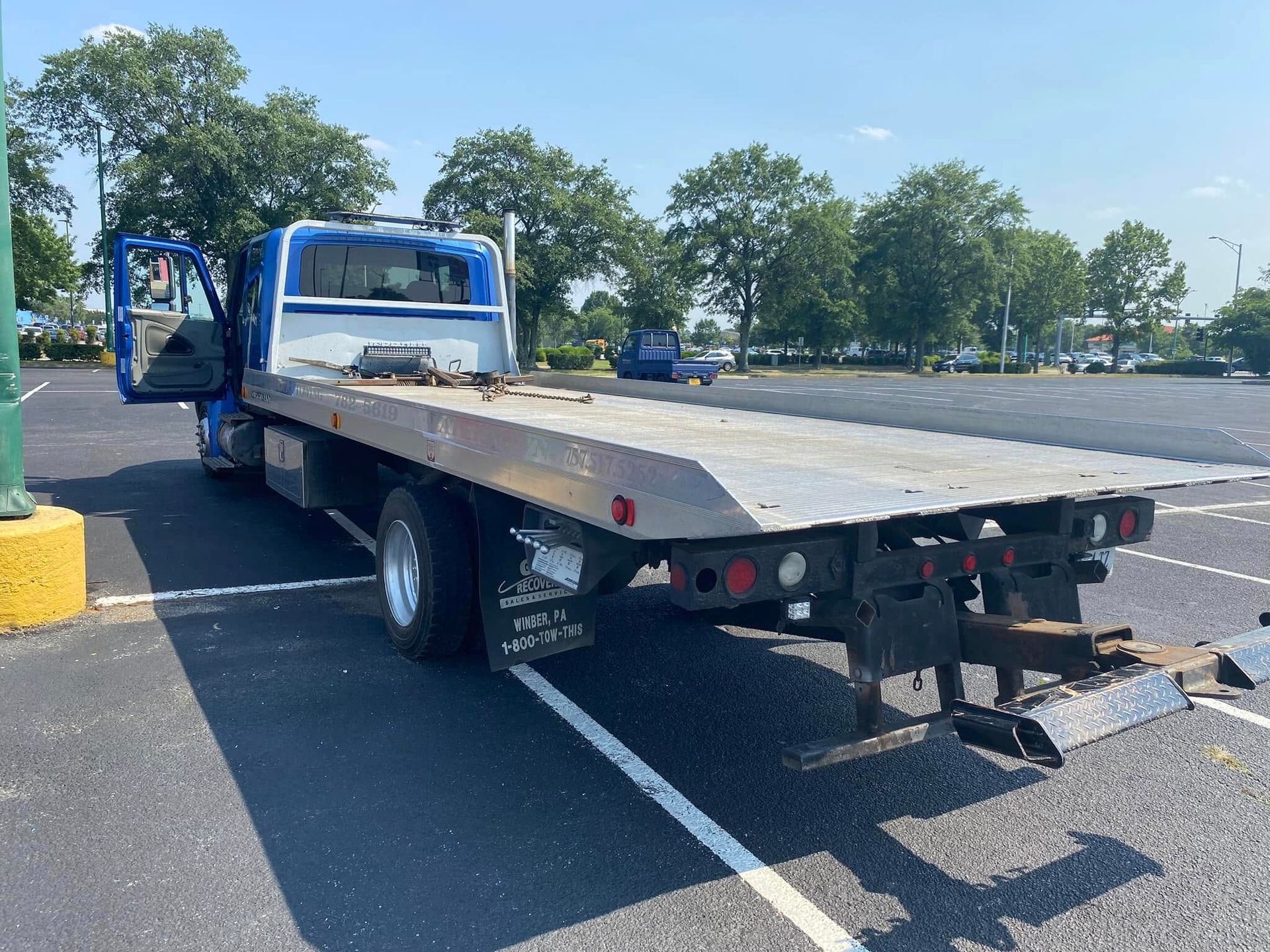 Flatbed tow truck parked in a sunny lot, with driver door open and rear towing hitch visible.