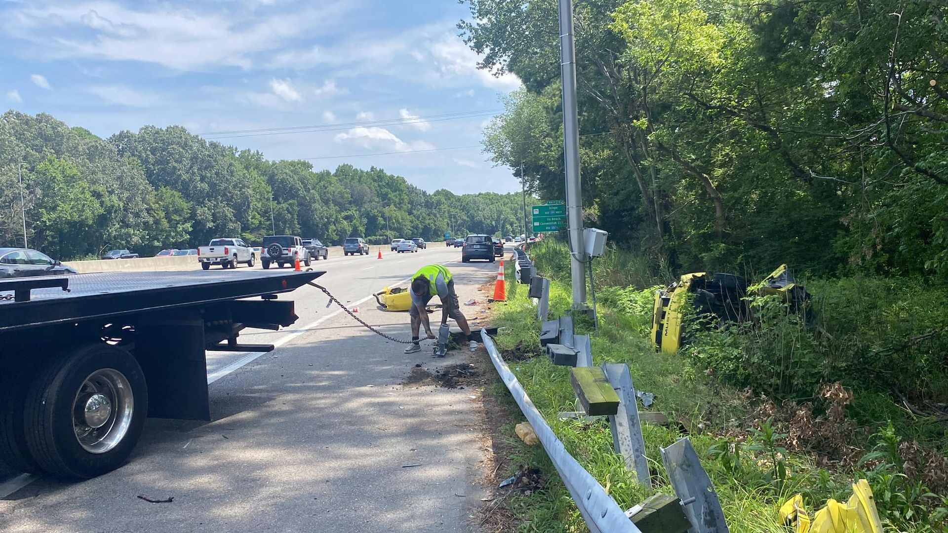 Tow truck workers on a highway shoulder beside a guardrail, loading a car in bright daylight