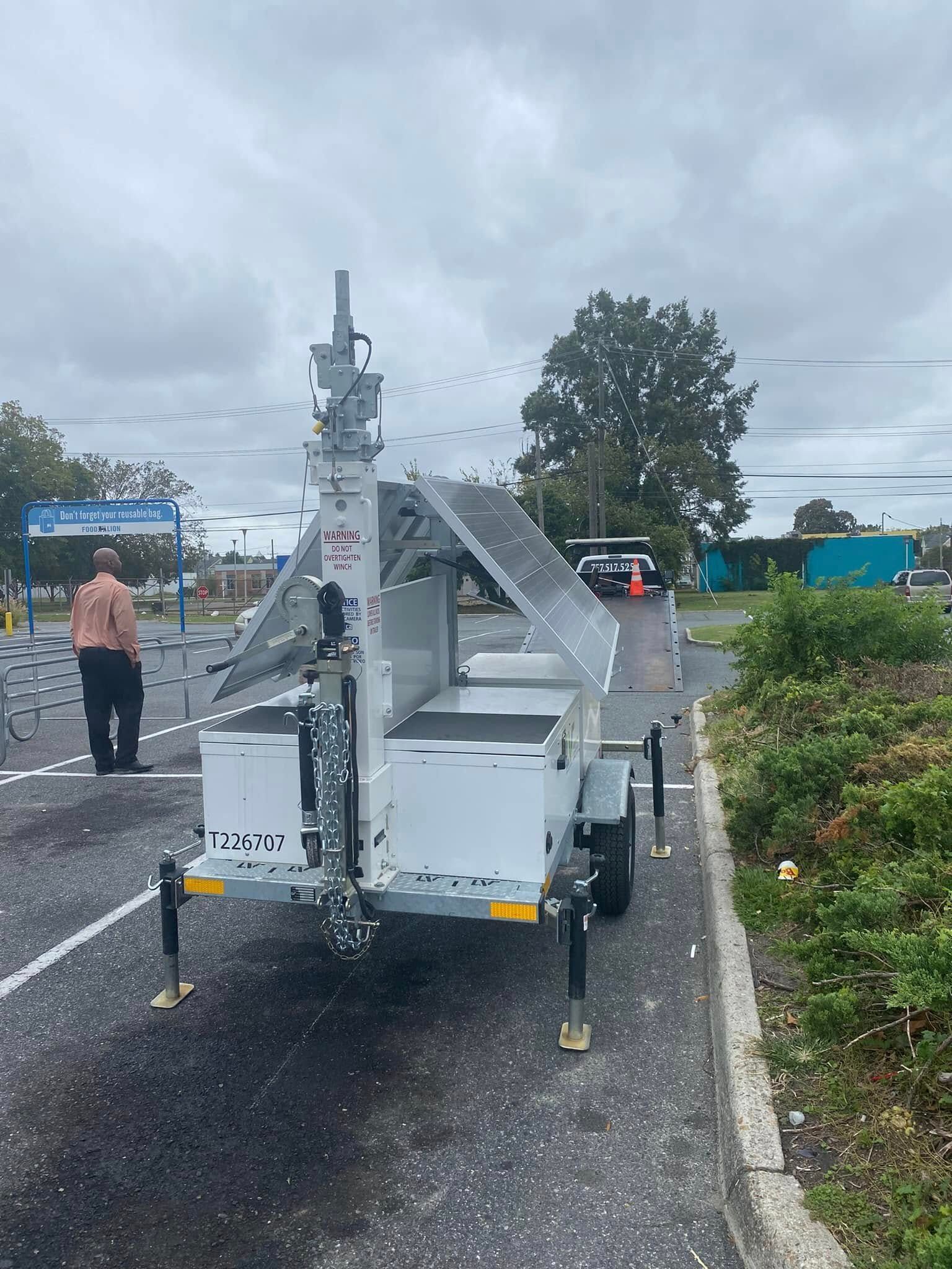 White utility trailer with raised equipment parked on a wet roadside under cloudy skies, with a person nearby