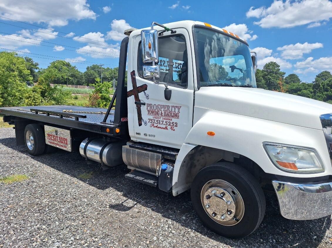 White flatbed tow truck parked on gravel under a blue sky with clouds