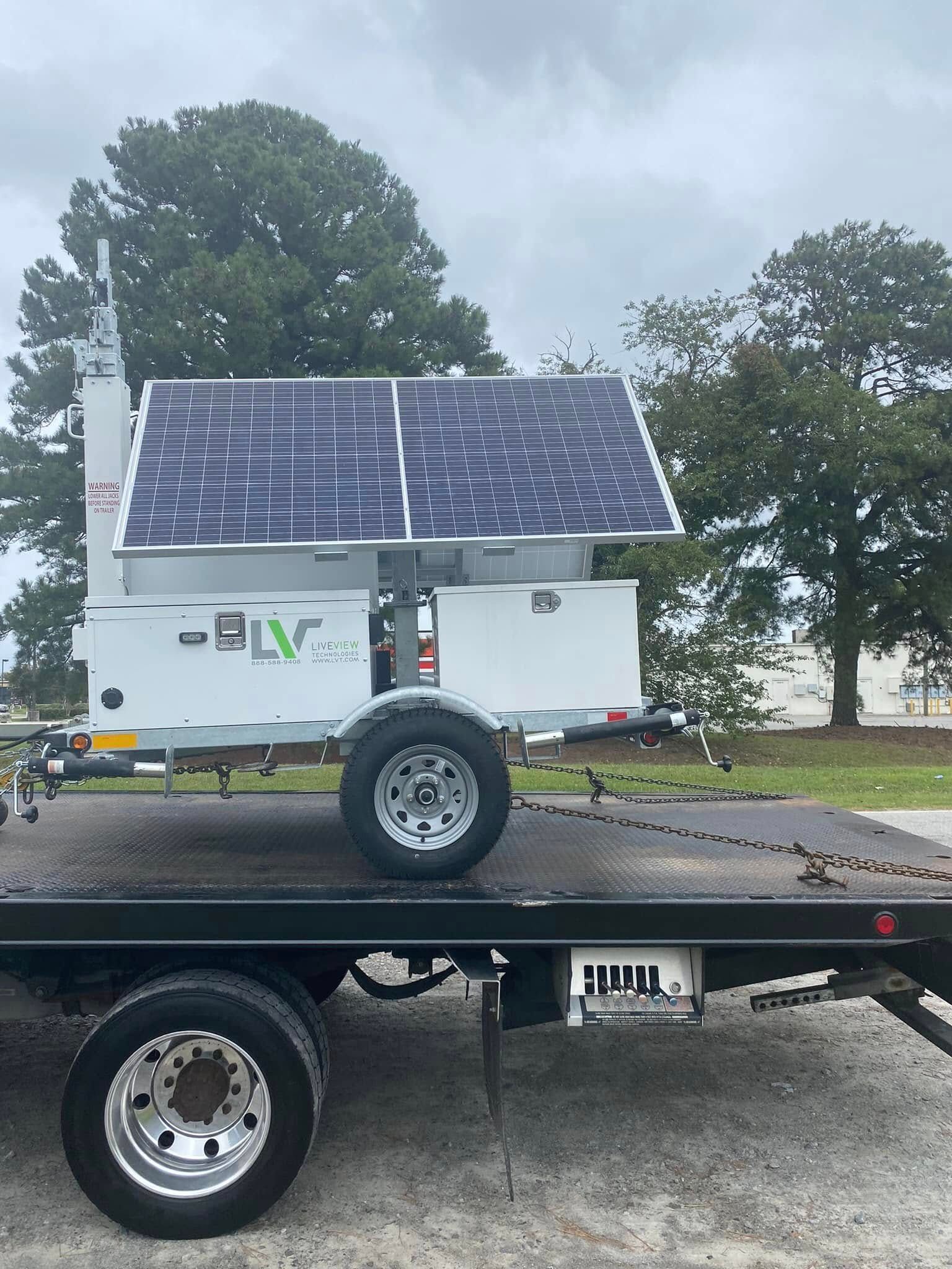 White solar trailer on a flatbed truck with large solar panels, parked outdoors under cloudy skies