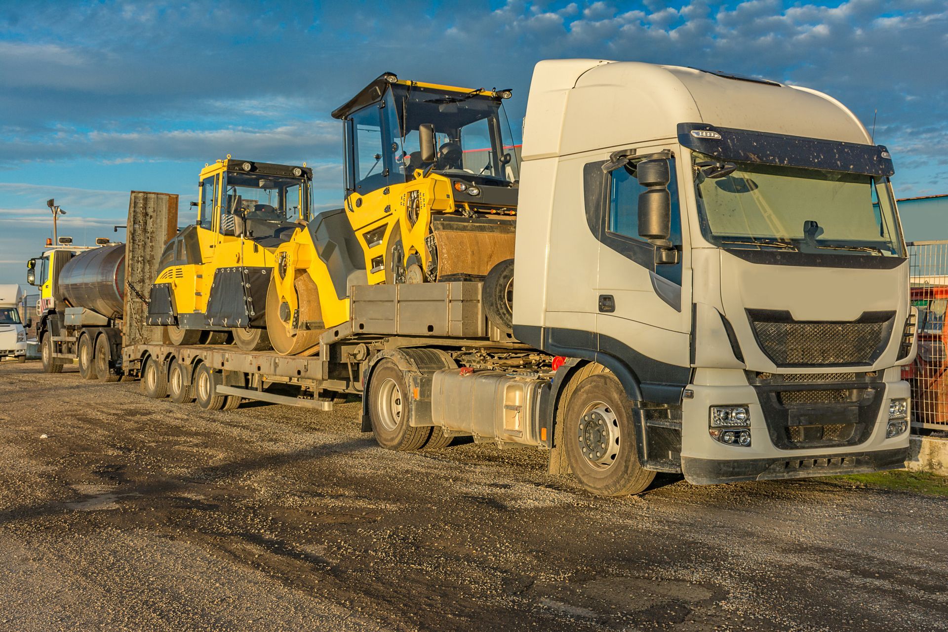 White semi-truck hauling yellow construction loaders on a flatbed trailer at a dirt worksite