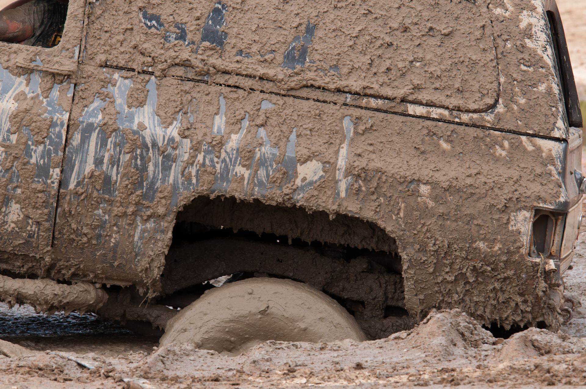 Mud-covered vehicle wheel stuck in thick dirt, with a large tire partially buried.