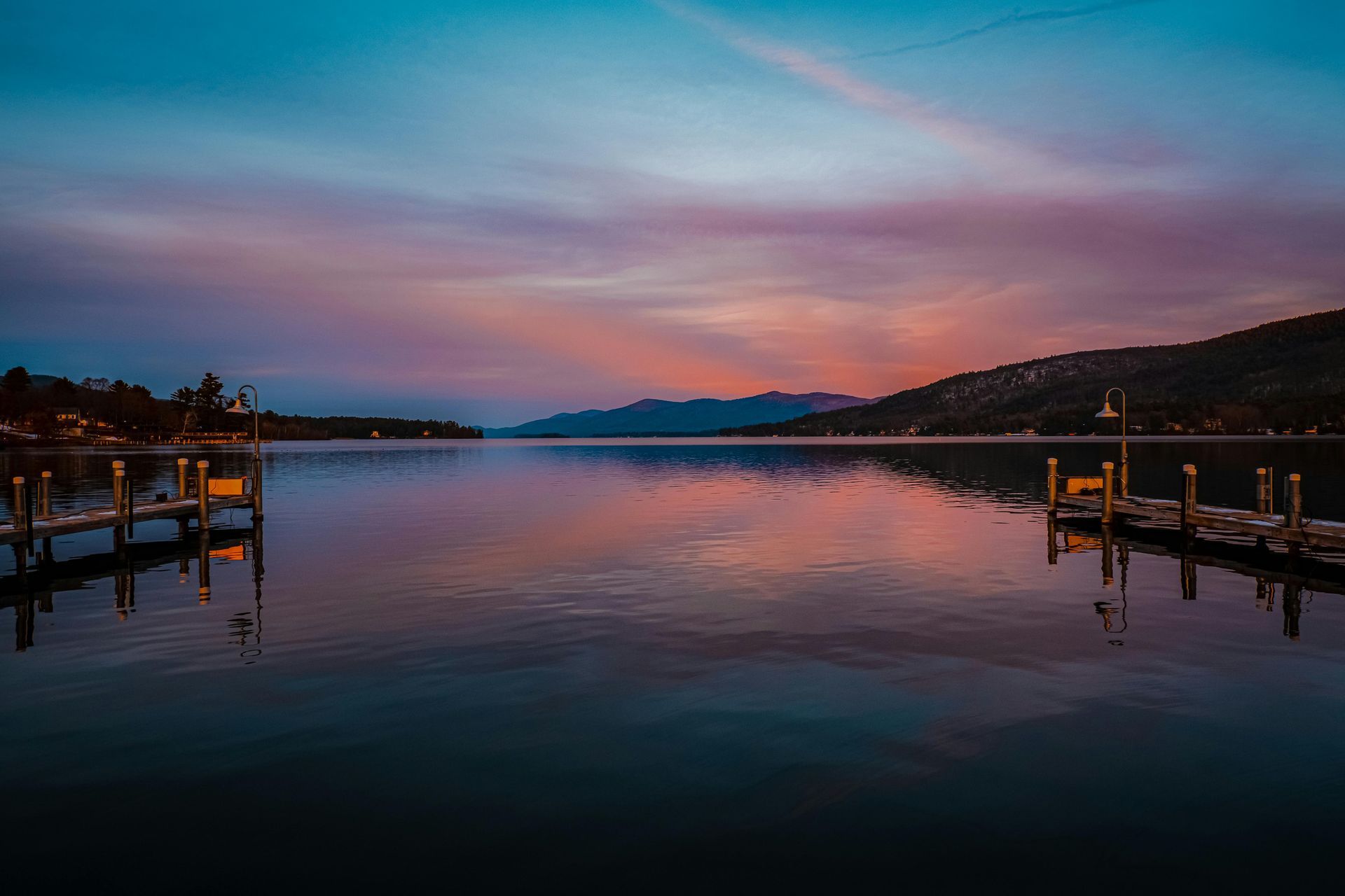 Lake at sunset, reflecting colorful sky. Wooden docks on each side, mountains in the distance.