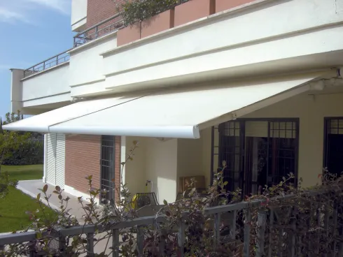 Tenda da sole bianca sopra il balcone di una casa in mattoni con ringhiera nera e piante da giardino davanti.