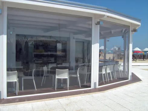 Caffè sul lungomare con pareti in vetro e sedie bianche sotto un patio coperto.
