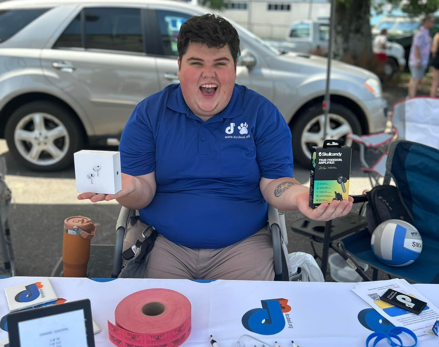 A person in a wheelchair smiles while holding an Apple AirPods box and a Belkin wireless charger at an outdoor booth.