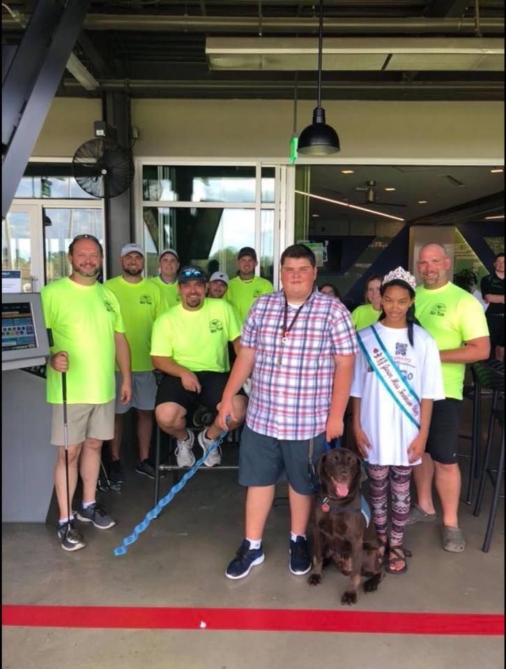 A group of people wearing neon yellow shirts and one in a plaid shirt pose with a dog at an indoor facility.