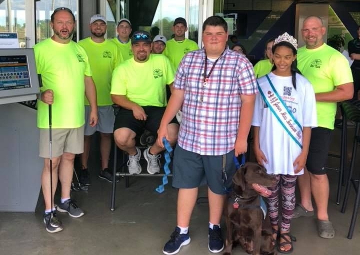 A group of people wearing neon yellow shirts stands with a girl wearing a sash and tiara, and a dog, at an indoor event.