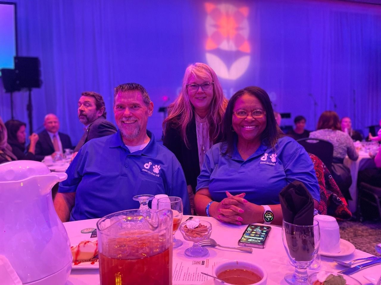 Three people sit at a round table at an event with purple lighting, smiling and looking toward the camera.