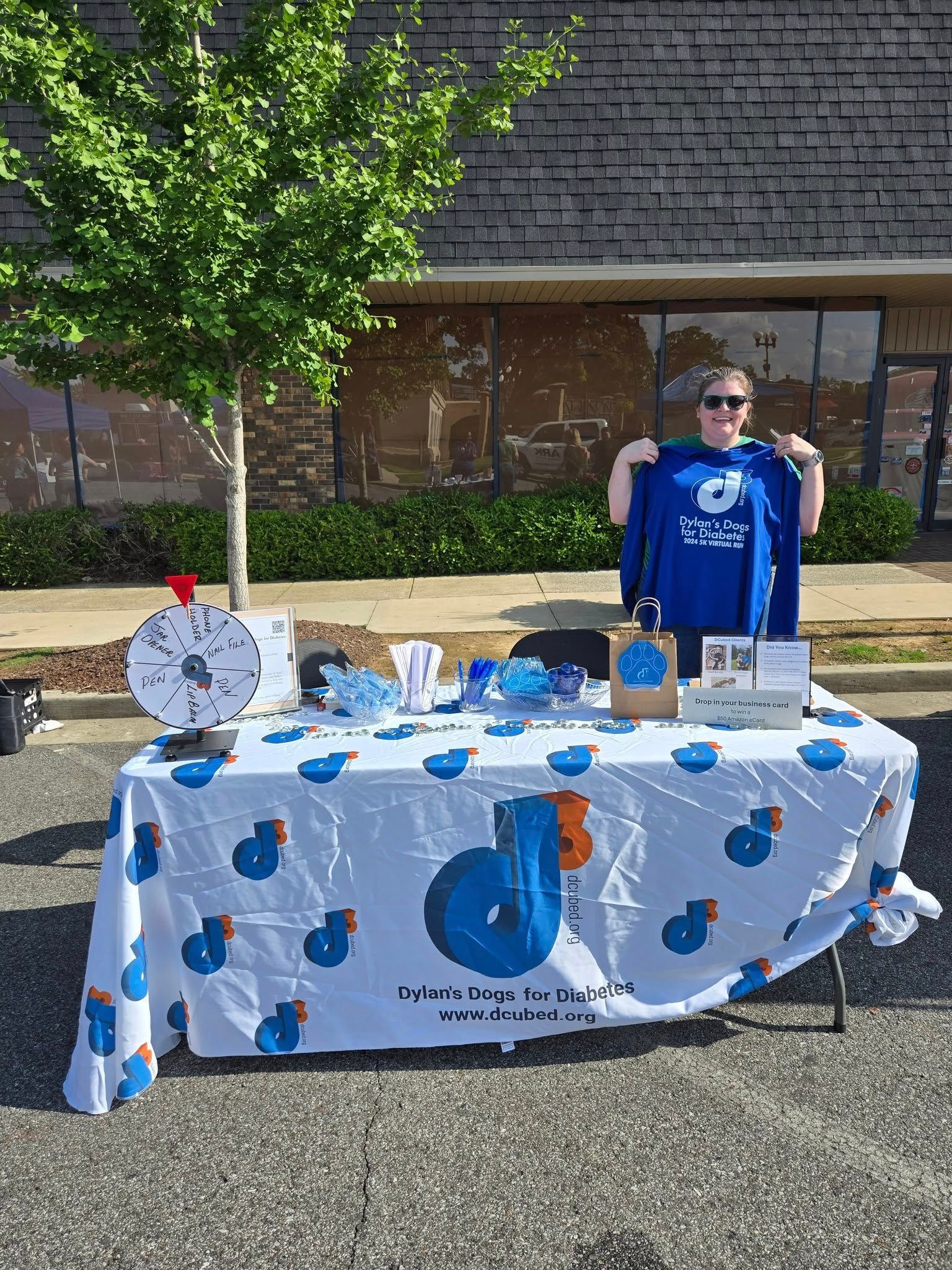 A person stands behind a branded booth table with a prize wheel, outdoors on a sunny day.