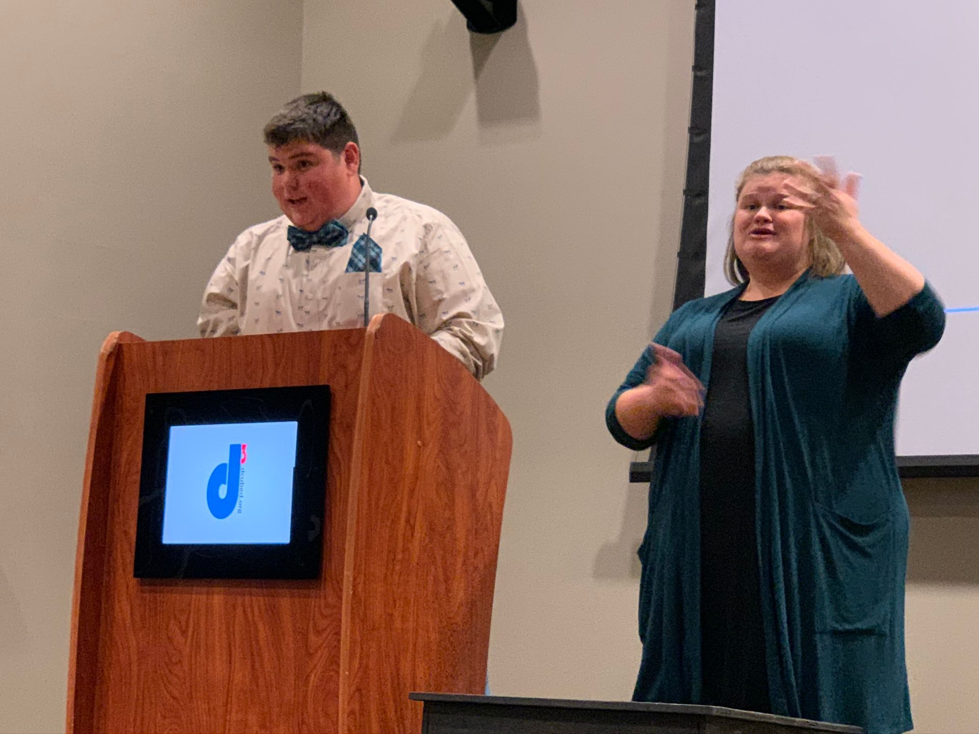 A speaker in a bow tie stands at a podium with a logo while an interpreter signs in front of a screen.