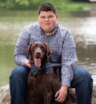 A person in a blue checkered shirt sits with a brown Labrador wearing a polka-dot bow tie by a lake.