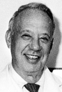 A man in a white coat and tie is smiling in a black and white photo.