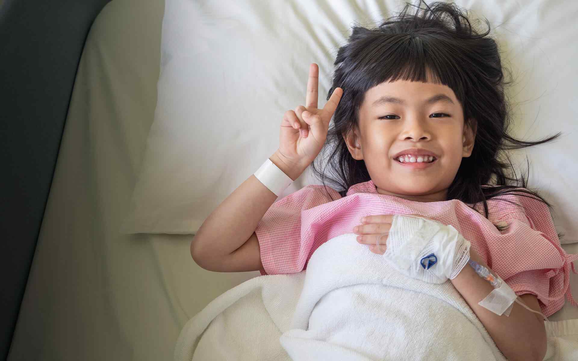 Smiling child lying on a bed, making a peace sign and holding a white stuffed rabbit