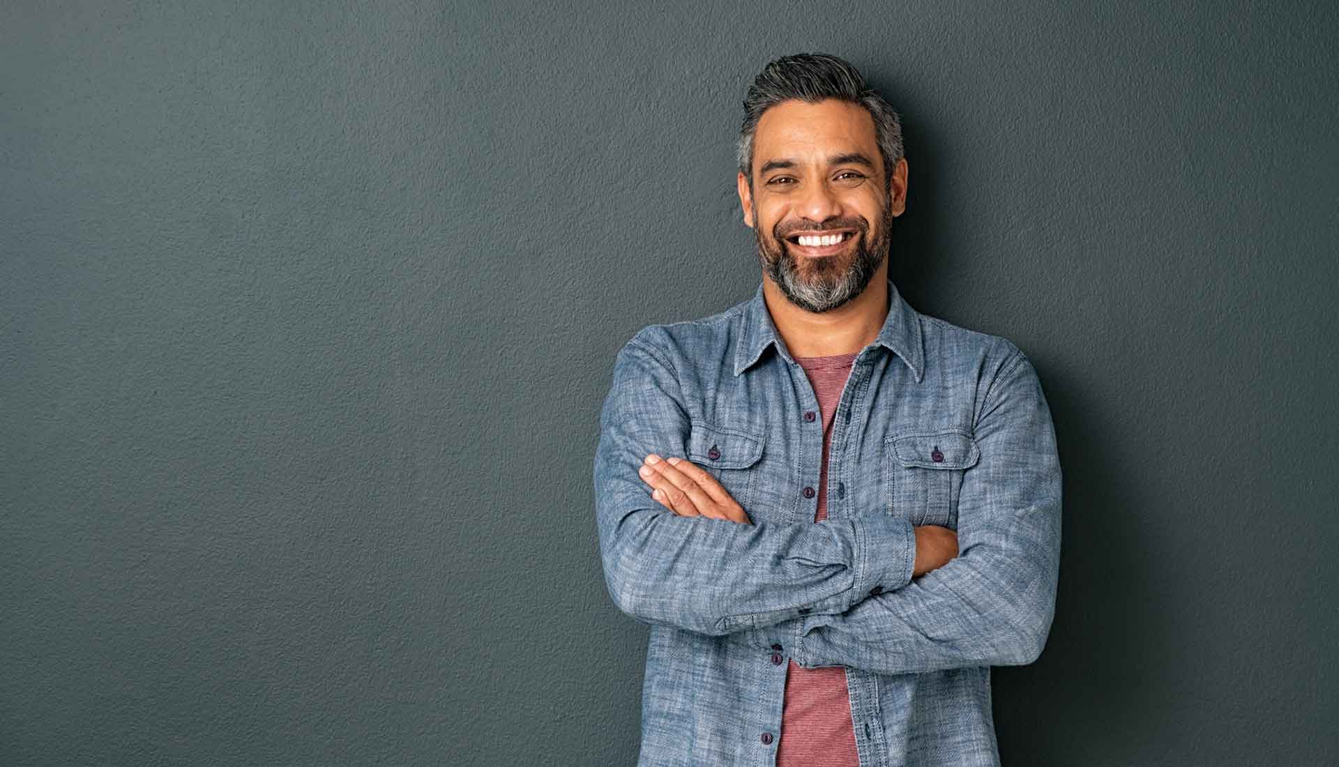 Smiling man with crossed arms, wearing a blue shirt against a dark gray wall