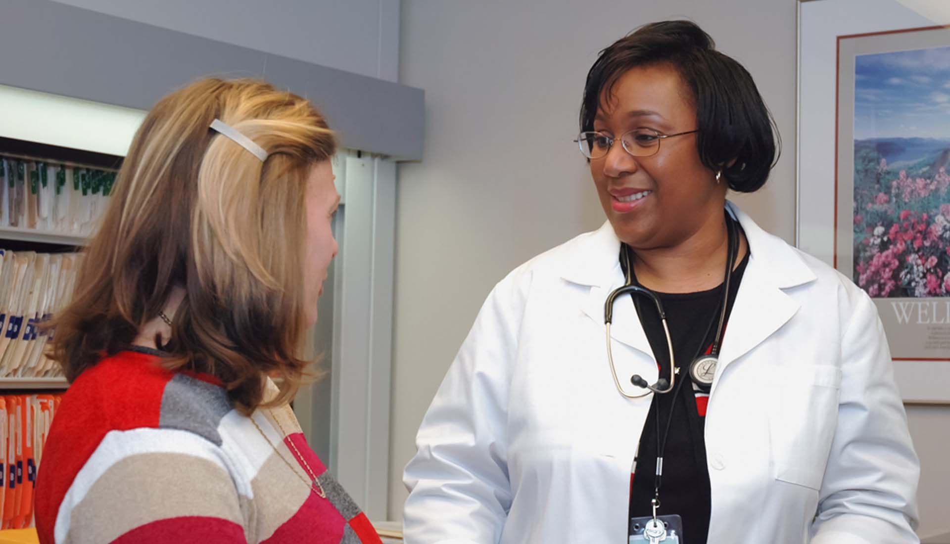 Doctor speaking with a patient in a clinic office, both smiling, with medical posters in the background.