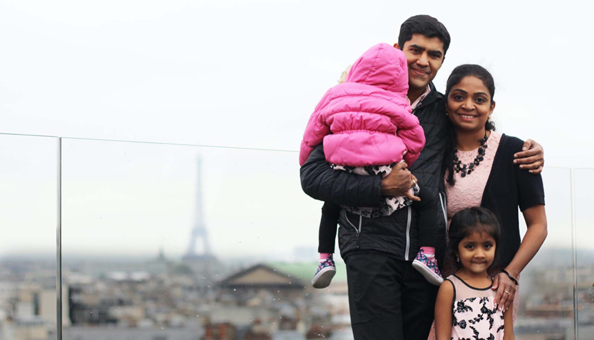 Family of four posing on a rooftop with the Eiffel Tower blurred in the background; one child is held in a pink jacket.