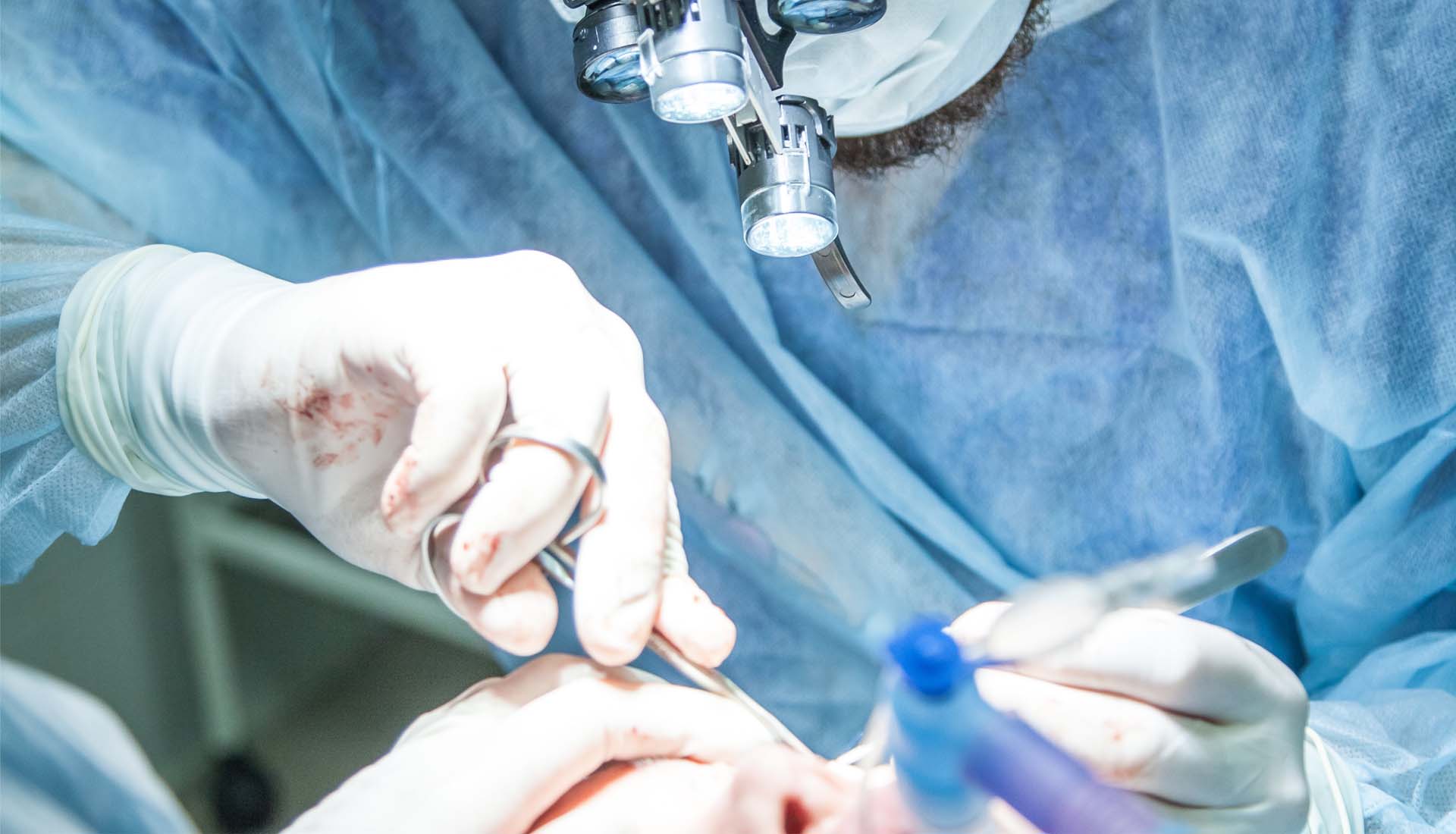Surgeons in blue scrubs performing an operation with gloved hands and surgical instruments under an operating microscope
