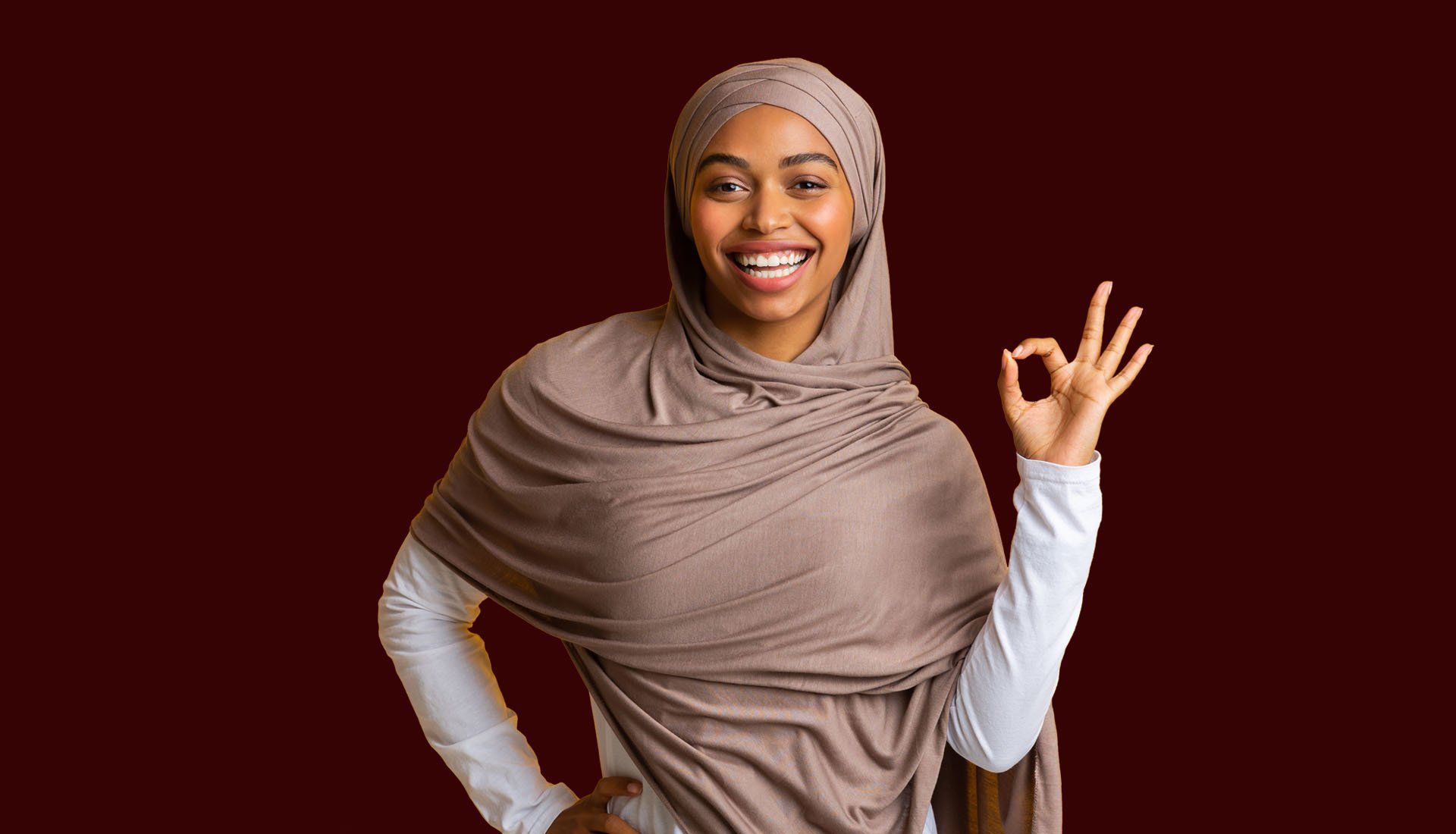 Smiling person in a beige hijab and shawl, making an OK hand sign against a dark red background