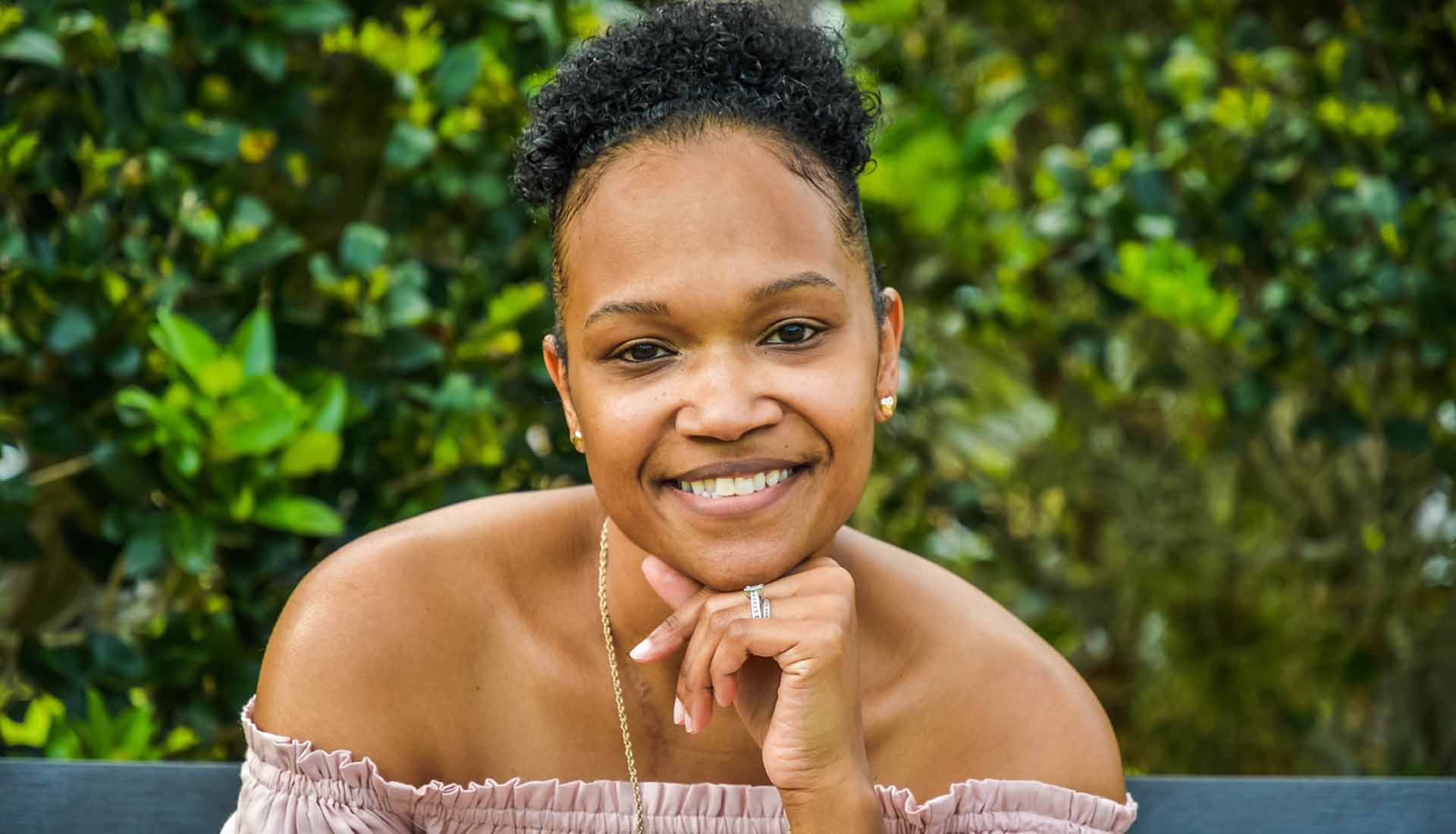 Smiling person leaning on a ledge outdoors with green foliage in the background