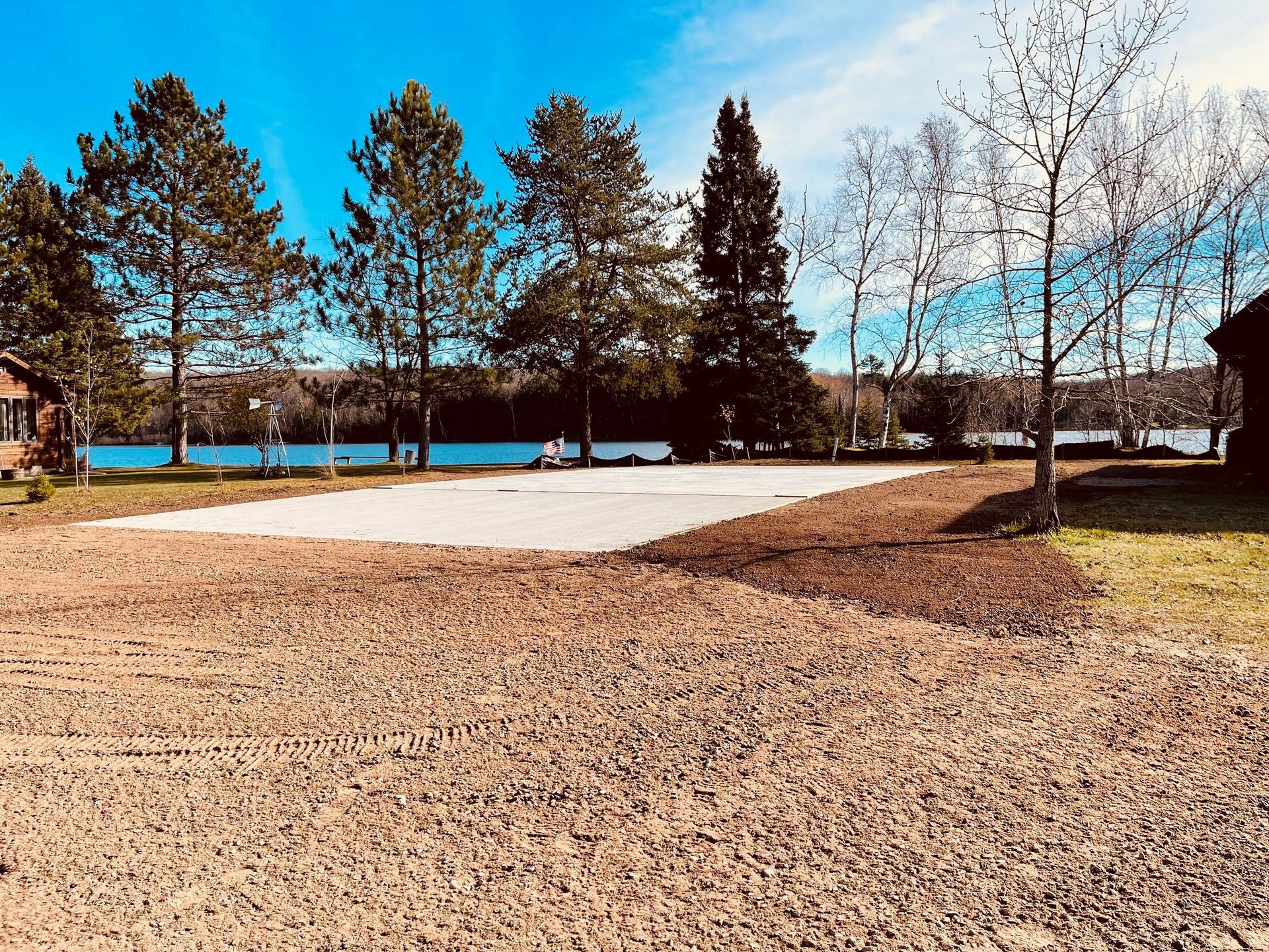 A rectangular concrete pad sits on a gravel lot near a lake, surrounded by evergreen trees under a bright blue sky.