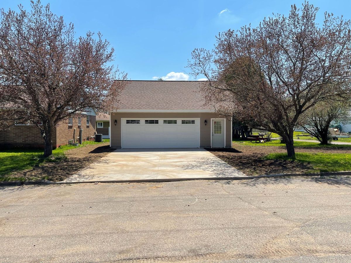 A beige detached garage with a white door and a side entry door, centered between two trees under a clear blue sky.