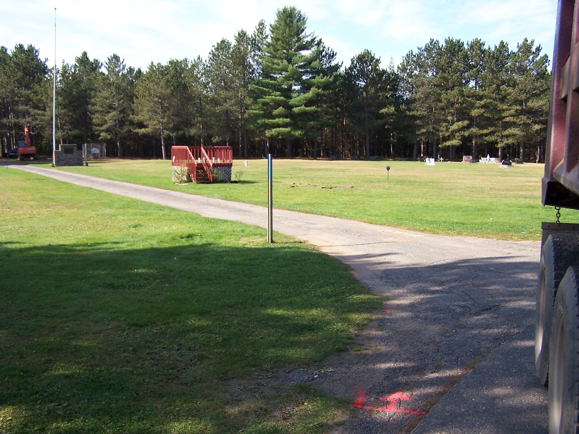 A gravel driveway leads through a grassy field toward a forest, with a red dumpster visible near the treeline.