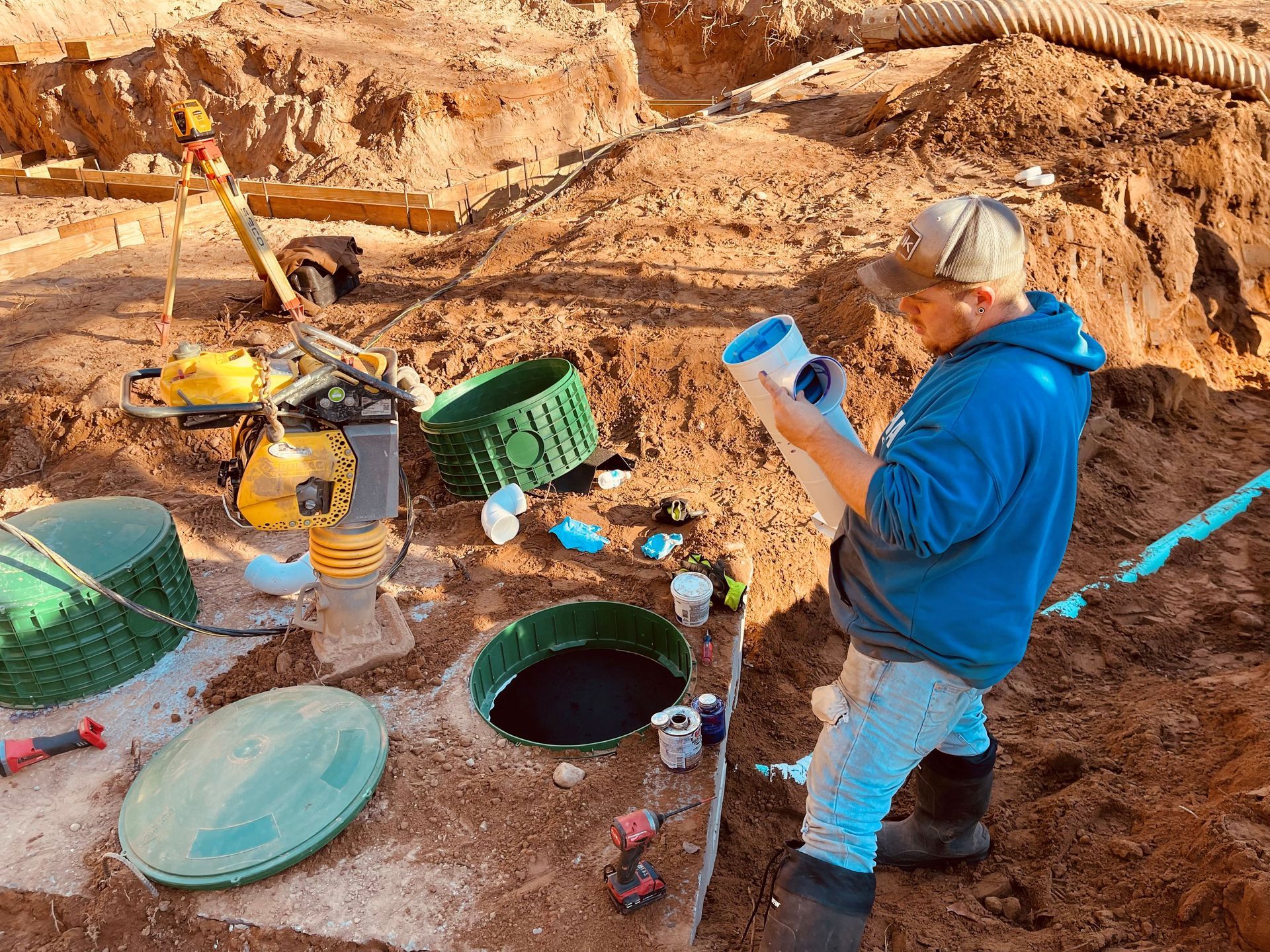 A worker in a blue hoodie stands in an excavation pit, inspecting a white PVC pipe near green septic tank risers. A worker in a blue hoodie stands in an excavation pit, inspecting a white PVC pipe near green septic tank risers.