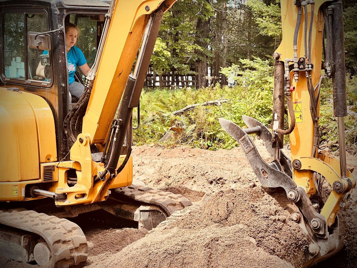 A person in a blue shirt operating a yellow excavator in a dirt-filled, wooded area.