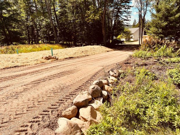 A dirt driveway with tire tracks, bordered by a stone wall on the right and a grassy bank on the left, leading to a house.