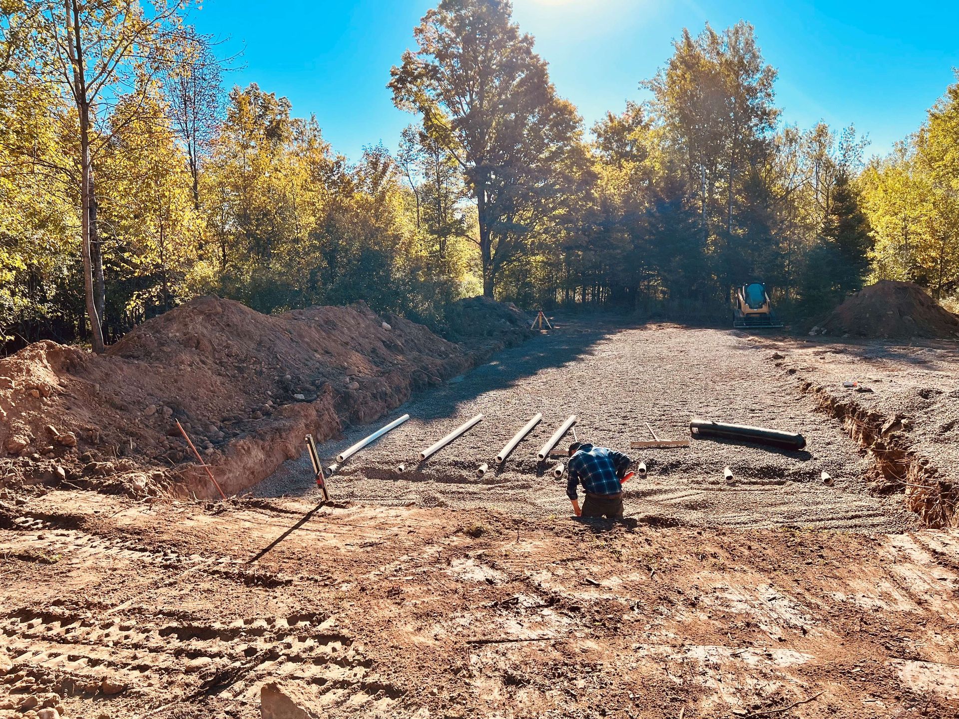 A person works in a muddy construction site with trenches, gravel ground, and surrounding trees under a bright blue sky.
